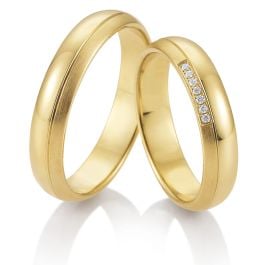 Zwei goldene Trauringe, einer davon mit einer Reihe kleiner Diamanten, auf einer spiegelnden weißen Oberfläche - perfekt für moderne Bräute in Aalen, die zeitlose Eleganz suchen.