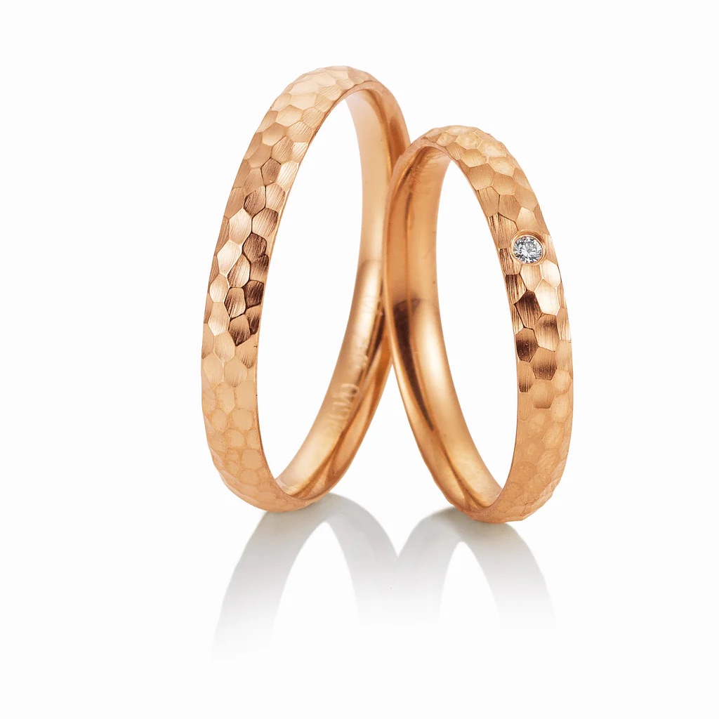 Zwei Ringe aus strukturiertem Roségold, einer davon mit einem kleinen Diamanten, stehen aufrecht auf weißem Grund - perfekt für moderne Bräute, die auf der Suche nach einzigartiger Eleganz sind, um exklusive Brautmode zu ergänzen.