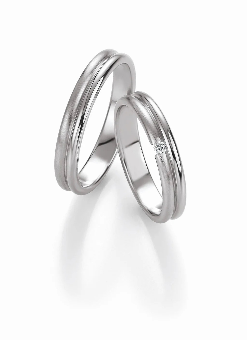 Zwei silberne Eheringe, einer mit einem kleinen Diamanten, auf weissem Grund - das perfekte Symbol für moderne Bräute in der Schweiz, passend zur exklusiven Brautmode.