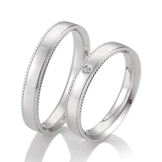 Zwei silberne Trauringe, einer mit einem kleinen Diamanten, beide mit Perlenrand - eine zeitlose Wahl für Paare in der Schweiz, passend zur Eleganz moderner Brautkleider.