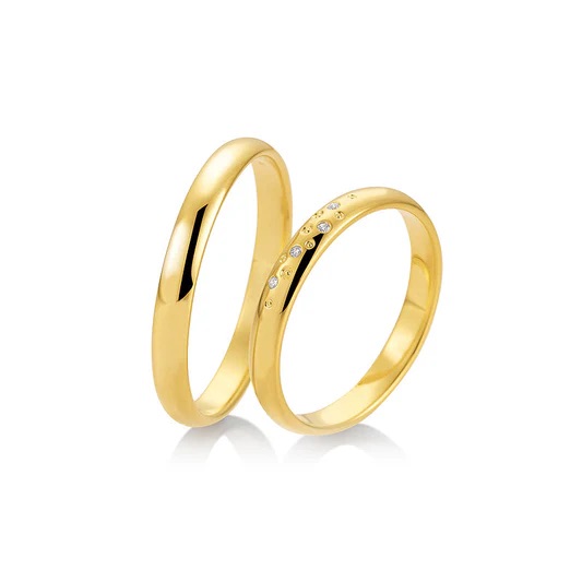 Zwei goldene Ringe - einer schlicht, einer mit kleinen eingelassenen Diamanten - glänzen auf weißem Grund, spiegeln die Eleganz der exklusiven Stuttgarter Brautmode wider und inspirieren zum Träumen von modernen Brautkleidern.