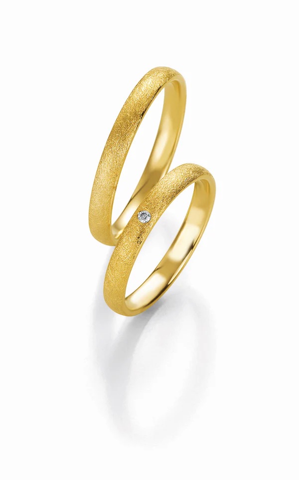 Zwei goldene Eheringe, einer davon mit einem kleinen Diamanten, auf weißem Hintergrund mit weichem Schatten - perfekt für moderne Bräute in der Schweiz.