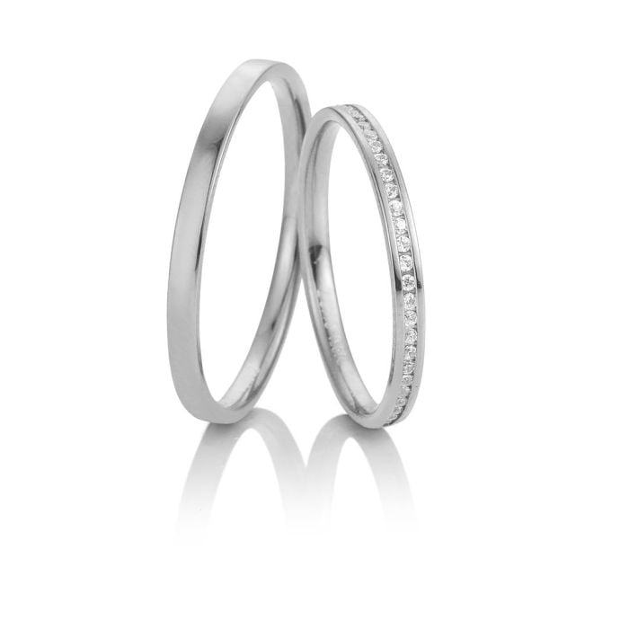Zwei silberne Eheringe, einer schlicht und einer mit kleinen Diamanten, stehen aufrecht auf einer weißen Fläche - perfekte Symbole für moderne Bräute, inspiriert von exklusiver Brautmode.