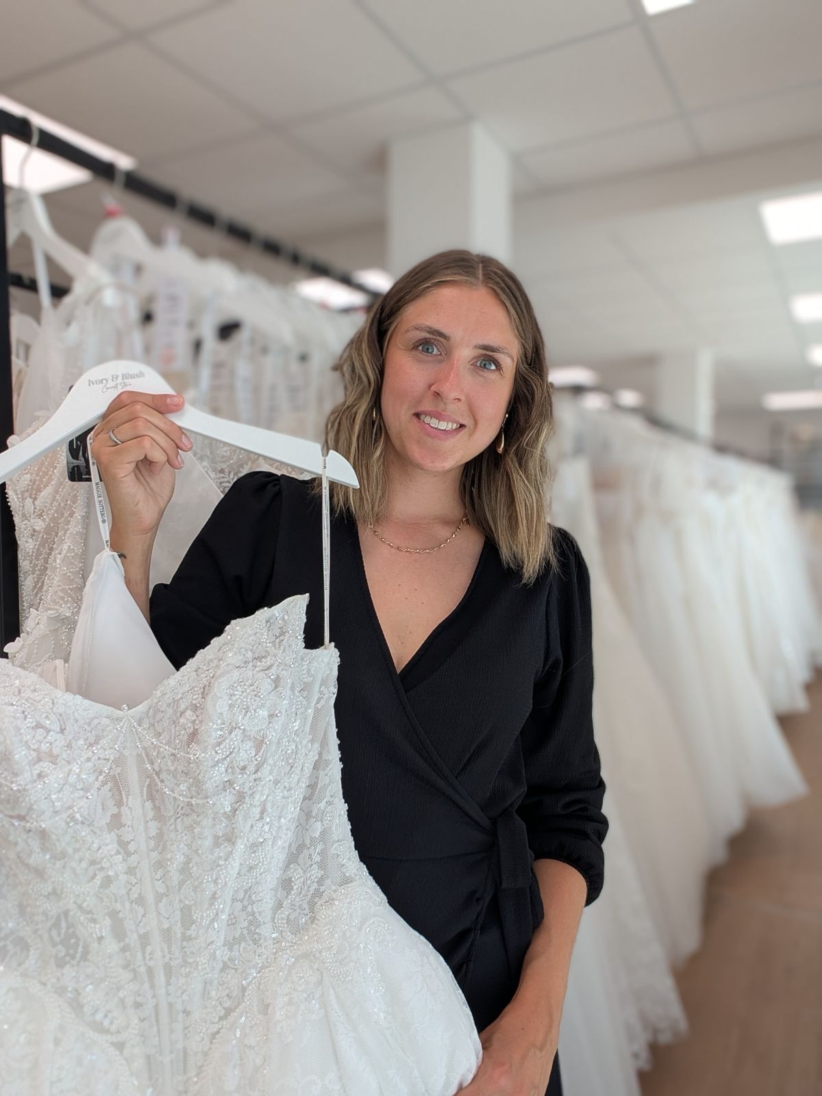 Eine Frau in schwarzer Kleidung hält in einem modernen Brautmodengeschäft ein weißes Spitzenhochzeitskleid auf einem Kleiderbügel in der Hand und präsentiert damit elegante Brautmode für die anspruchsvolle Braut.